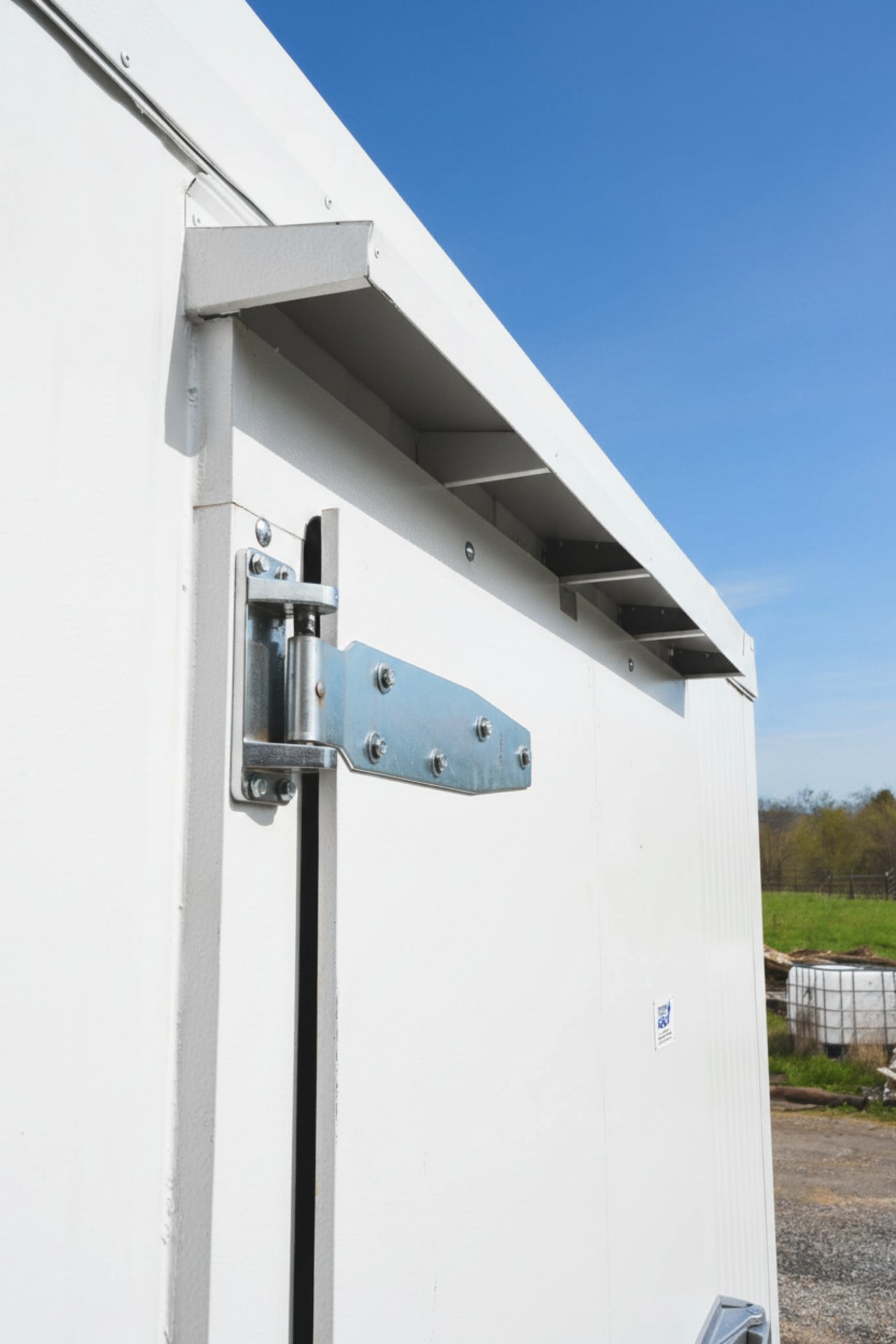 Heavy bright-metal hinge and the roof drip edge at the corner of the Berkshire Grown outdoor box, farmland behind it